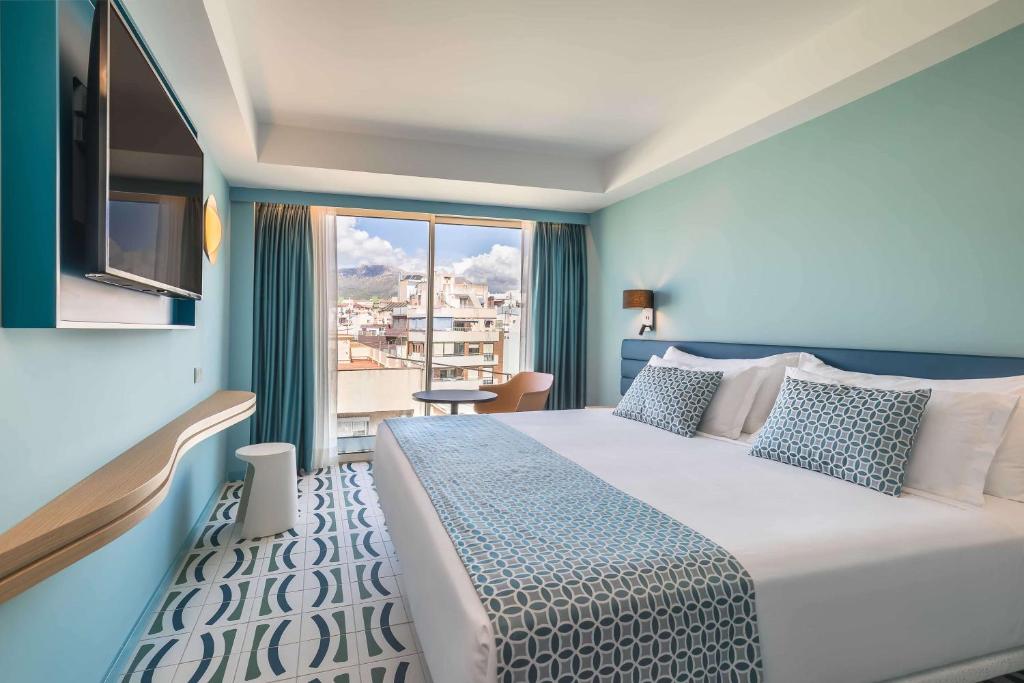 Mūsdienīga viesnīcas istaba ar gaišiem toņiem un lieliem logiem, kas sniedz skatu uz pilsētu. Istabā ir plaša gulta, televizors un stilīgs interjers ar akcentiem.