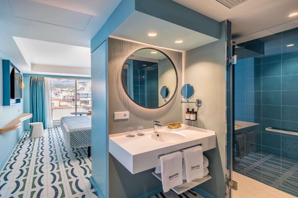 Mūsdienīgs viesnīcas numurs ar skatu uz pilsētu, kurā redzams elegants vannasistabas dizains ar apaļu spoguli un gaiši zilu apdari. Grīda ir izrotāta ar rakstainām flīzēm, radot stilīgu un komfortablu atmosfēru.