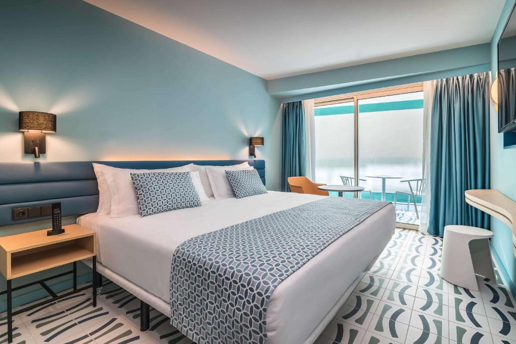 Mūsdienīga viesnīcas numura interjers ar gaišām krāsām, divguļamo gultu un balkonu, no kura paveras skats uz āra terasi.