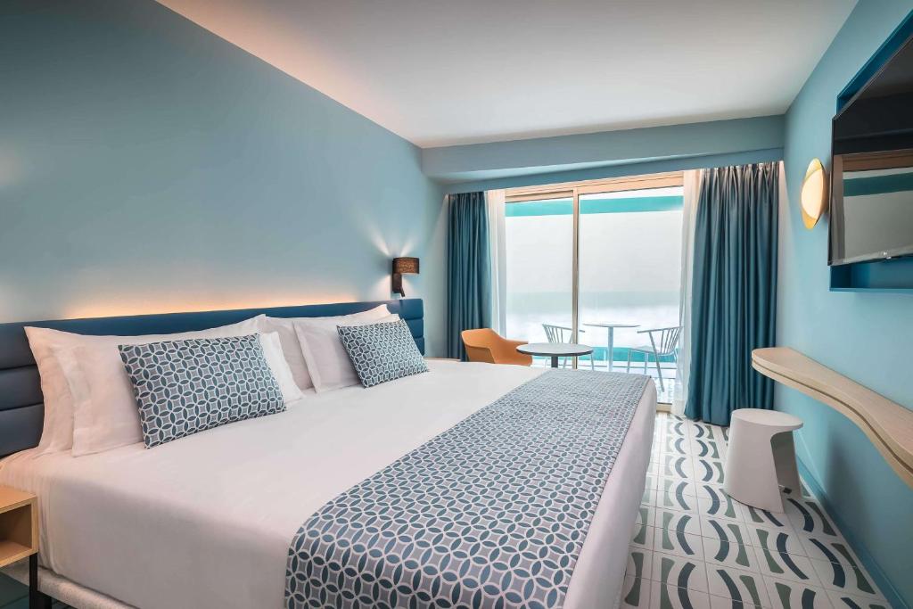 Mūsdienīgs viesnīcas numurs ar gaišām, zilu toņu sienām, plašu gultu un dekoratīvām spilveniem. Logi aizved uz balkonu ar skatu uz ārpusi, radot relaksējošu atmosfēru.