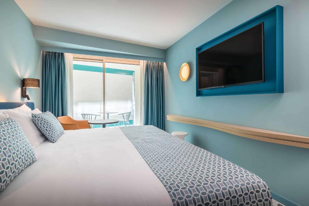 Mūsdienīga viesnīcas istaba ar gaišu zilo krāsu, plašu gultu un televizoru uz sienas. Logi ar aizkariem un balkona mēbeles nodrošina patīkamu skatu un gaismu.