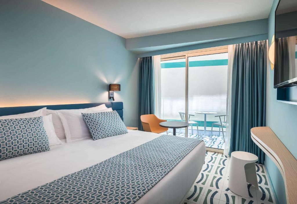 Moderna viesnīcas numuriņa interjers ar gaiši zilu sienu, ērtu gultu un stilīgiem aksesuāriem. Numurā ir balkons ar galdu un krēsliem, kas nodrošina patīkamu skatu.