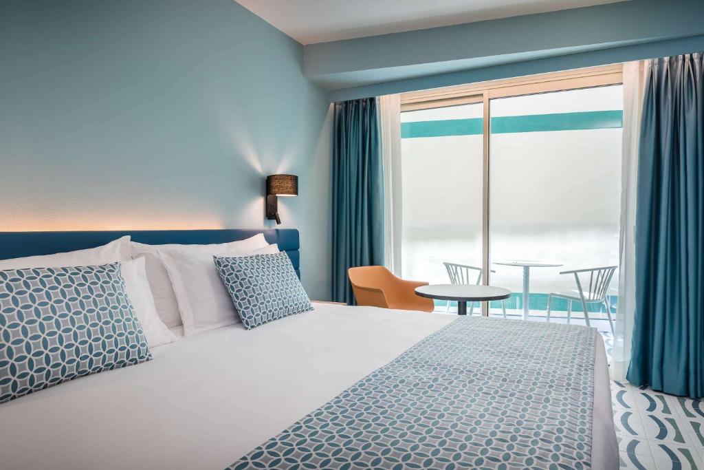 Mūsdienīgs viesnīcas numurs ar gaiši zilu sienu, ērtu gultu un dekoratīvām spilveniem. Numurā ir pieejama balkona zona ar galdu un krēsliem.