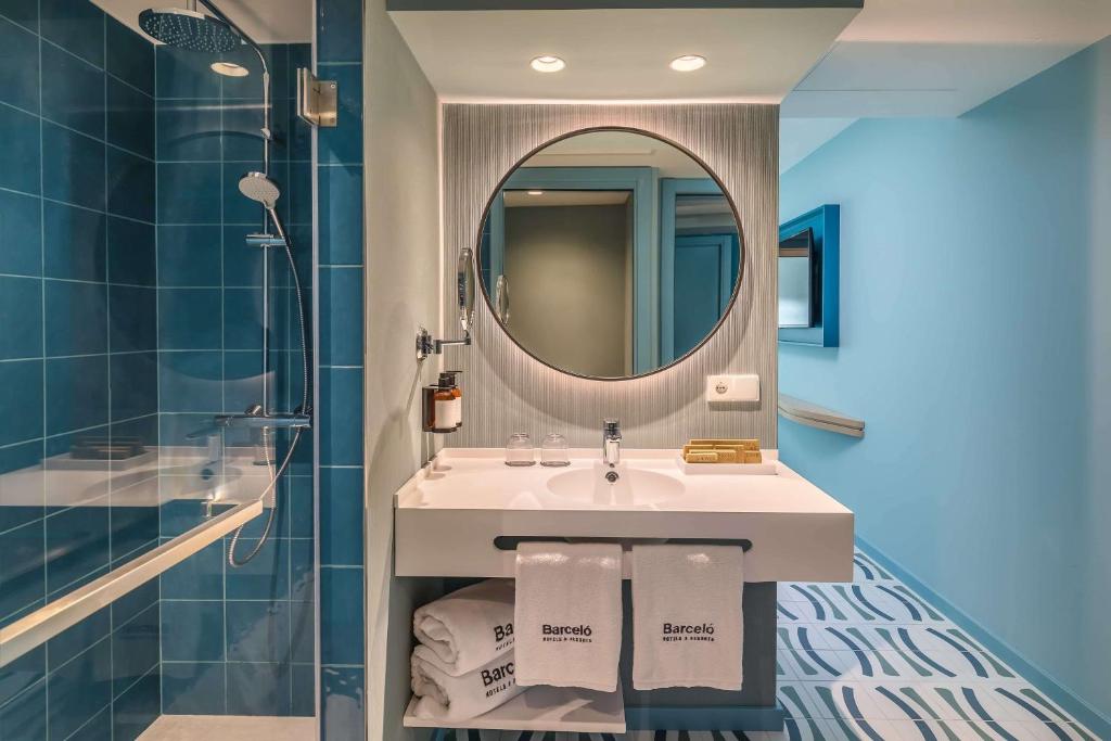 Mūsdienīgs vannas istabas dizains ar zilu flīžu apdari, apaļu spoguli un stilīgu izlietni. Vannas istabā redzami arī dvieļi un moderns dušas stūris.