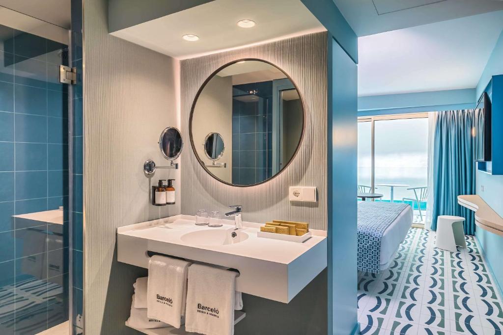 Mūsdienīgs viesnīcas numura vannas istabas interjers ar zilām flīzēm, apaļu spoguli un elegantu izkārtojumu. Gaismas akcenti un stilīga mājas dekorācija rada patīkamu atmosfēru.