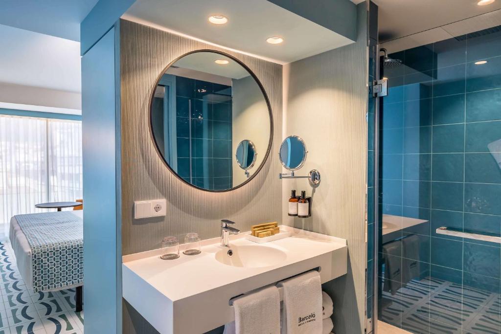 Mūsdienīga vannas istaba ar apaļu spoguli un stilīgu izlietni, kurā redzami higiēnas piederumi un dušas kabīne ar stikla durvīm. Sienas ir apšūtas ar zaļām flīzēm, radot svaigu un relaksējošu atmosfēru.