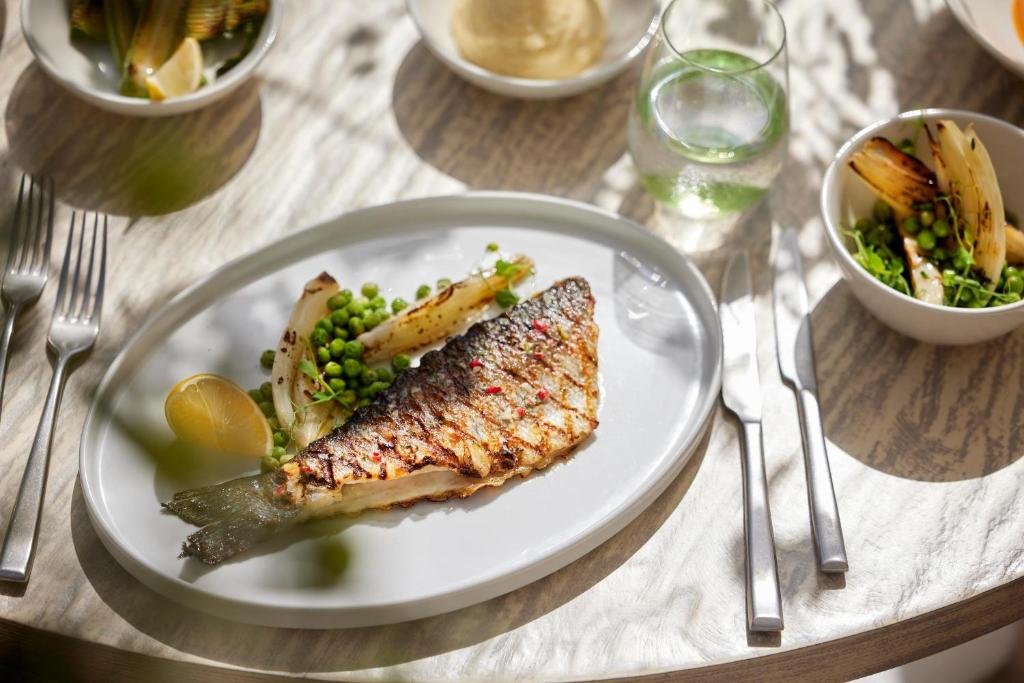 Grilēts zivs fileja ar zaļajiem zirnīšiem un citronu, pasniegta ar grilētu dārzeņu piedevu. Blakus atrodas ūdens glāze un salāti.