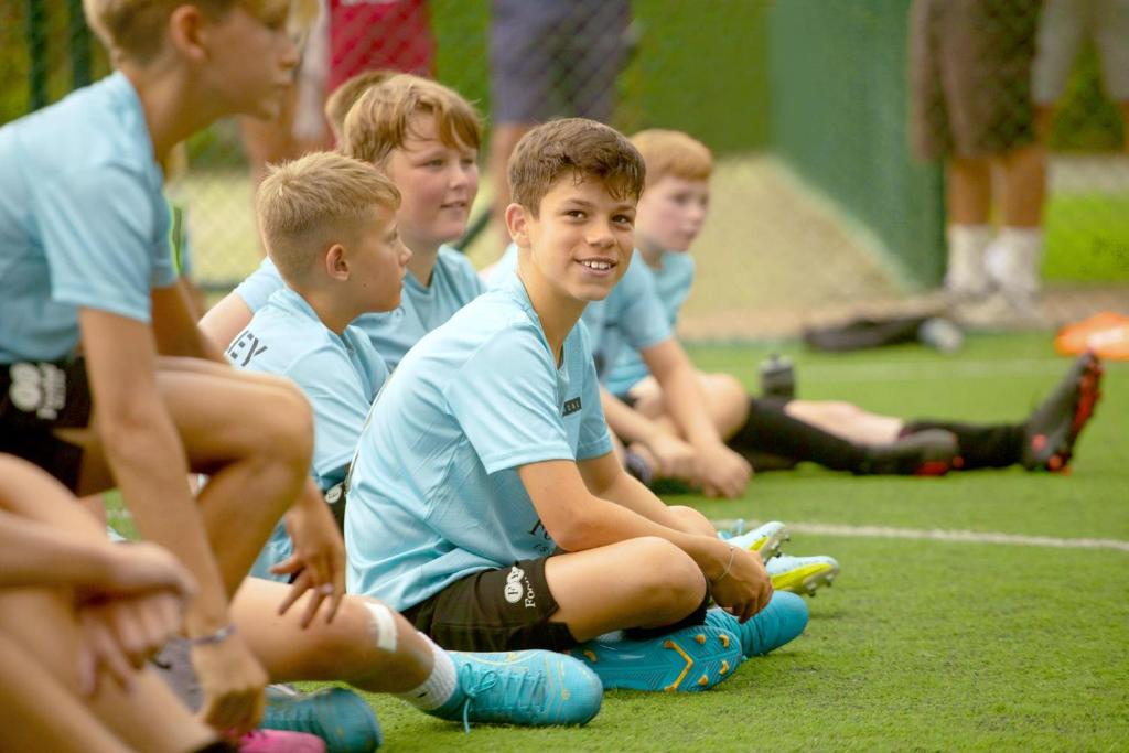 Jauni futbolisti sēž uz zāliena, gatavojoties treniņam. Priecīgs zēns ar brūniem matiem un zilām futbola kurpēm izsaka draudzīgu smaidu.