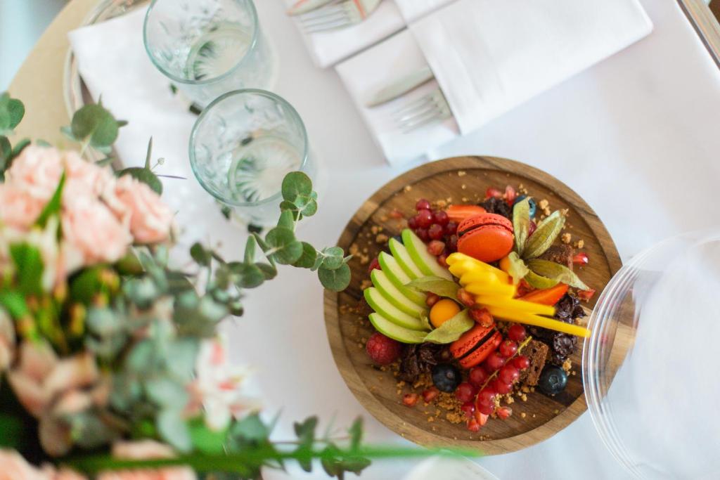 Krāšņa augļu un izstrādājumu kompozīcija koka traukā, apvienojot zaļās ābolus, ogas un makaronus, blakus ziedu pušķis un glāzes.