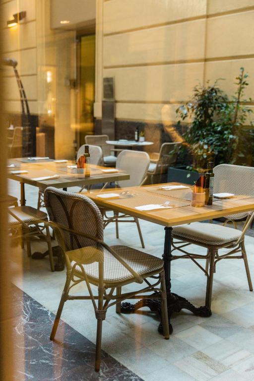 Elegants restorāna interjers ar koka galdiem un krēsliem, kas gaida viesus. Gaisma caur logu apgaismo telpu, radot patīkamu atmosfēru.