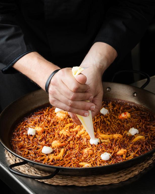 Šefpavārs dekorē ēdienu ar krējuma maisījumu, izmantojot konditorejas maisiņu, uzsverot krāsainos rīsus un garneles pannā.