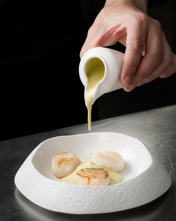 Šefpavārs ielej zaļo mērci uz trīs apceptām jūras veltēm, izmantojot elegantu balto trauku. 