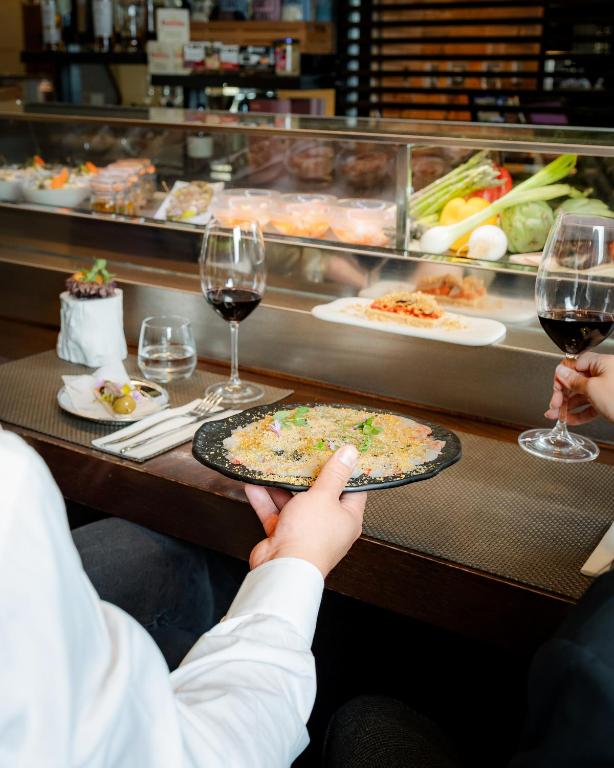 Elegants restorāna iestatījums ar viesi, kas tur šķīvi ar izsmalcinātu ēdienu, un glāzi sarkanvīna. Fonā redzami svaigi dārzeņi un citas maltītes.