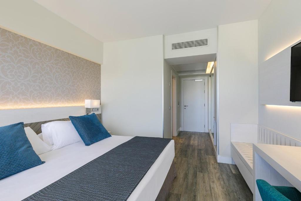 Mūsdienīga viesnīcas numura iekšskats ar gultu, zilām spilveniem un elegantu dizainu. Telpā ir arī televizors, koka grīdas un gaišas krāsas sienas.