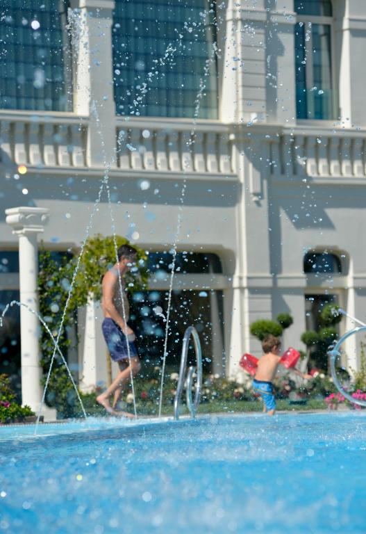 Bērns ar peldēšanas rokturiem spēlējas baseinā, kamēr pieaugušais staigā gar baseinu ar ūdens strūklām. Fonā redzams elegants ēku arhitektūras elements un zaļumi.