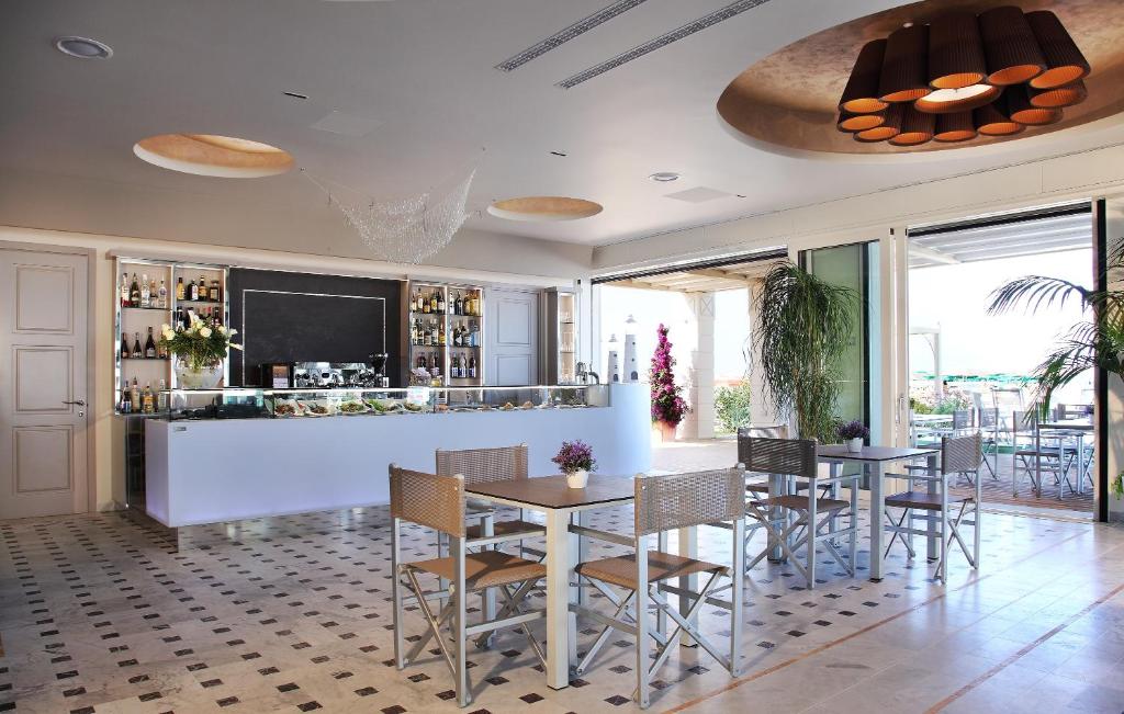 Moderns restorāns ar gaišu interjeru, dzīvām zaļām augiem un elegantu bāra zonu. Atvērtas durvis ved uz terasi ar skatu uz āru.