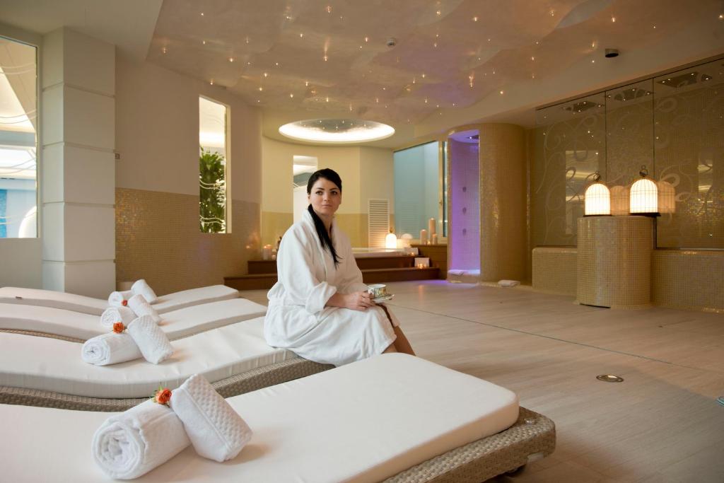 Skaista un relaksējoša spa vide ar komfortablām guļamvietām un mīkstām dvieļiem, kurā sieviete sēž, ģērbusies baltā halātā. Spa telpā ir mūsdienīgs interjers un mierīga atmosfēra.