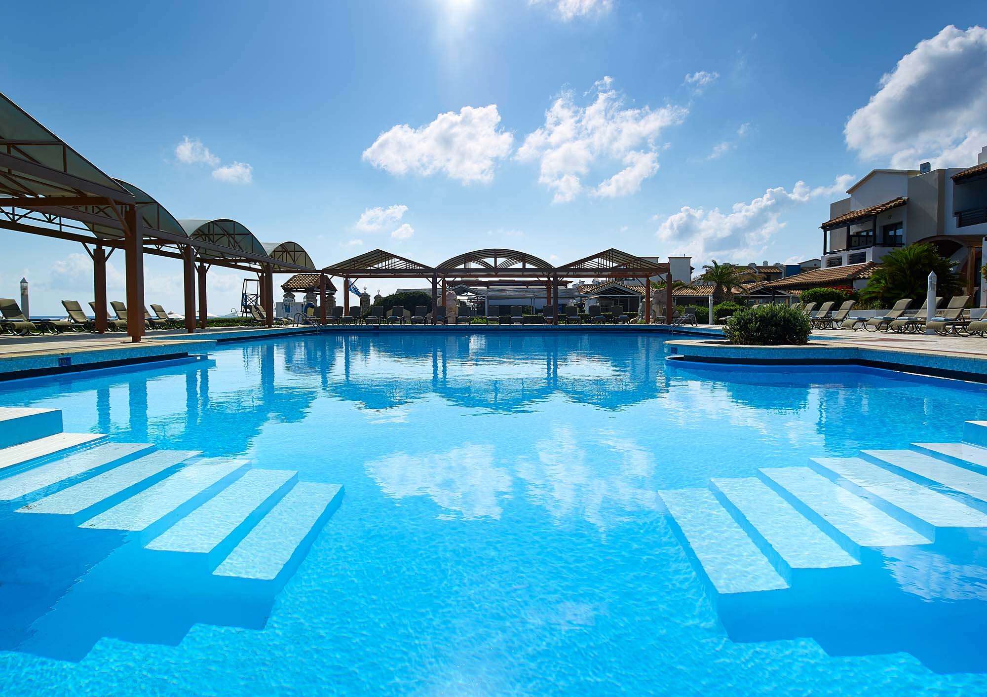 Liels, zila baseina skats ar atspulgām, saulains laiks un mājīga atpūtas zona zem nojumes. Ideāla vieta relaksācijai un brīvā laika pavadīšanai. panoramic view with beach and pools