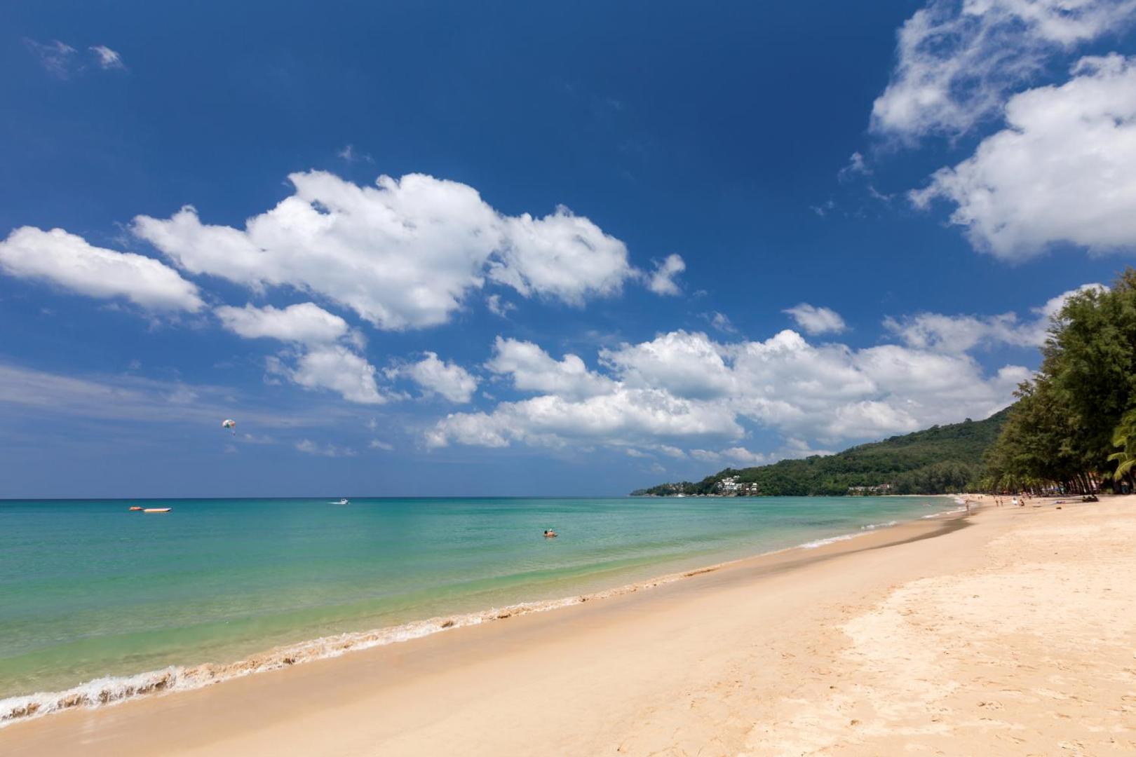 Sunwing Resort Kamala Beach pieguļošā smilšu pludmale ar dzidru tirkīza ūdeni un zaļiem kalniem tālumā Puketā