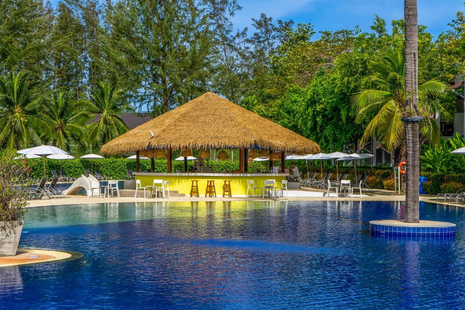 Sunwing Resort Kamala Beach āra baseins ar bāru zem salmu jumta Puketā, Taizemē — apkārtnē palmas, sauļošanās krēsli un atpūtas zona.