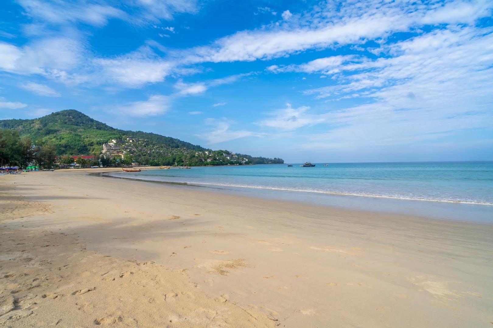 Sunwing Resort Kamala Beach panorāmas skats uz Kamala pludmali Puketā ar apkārtējiem zaļajiem kalniem un zilu debesu fonu.