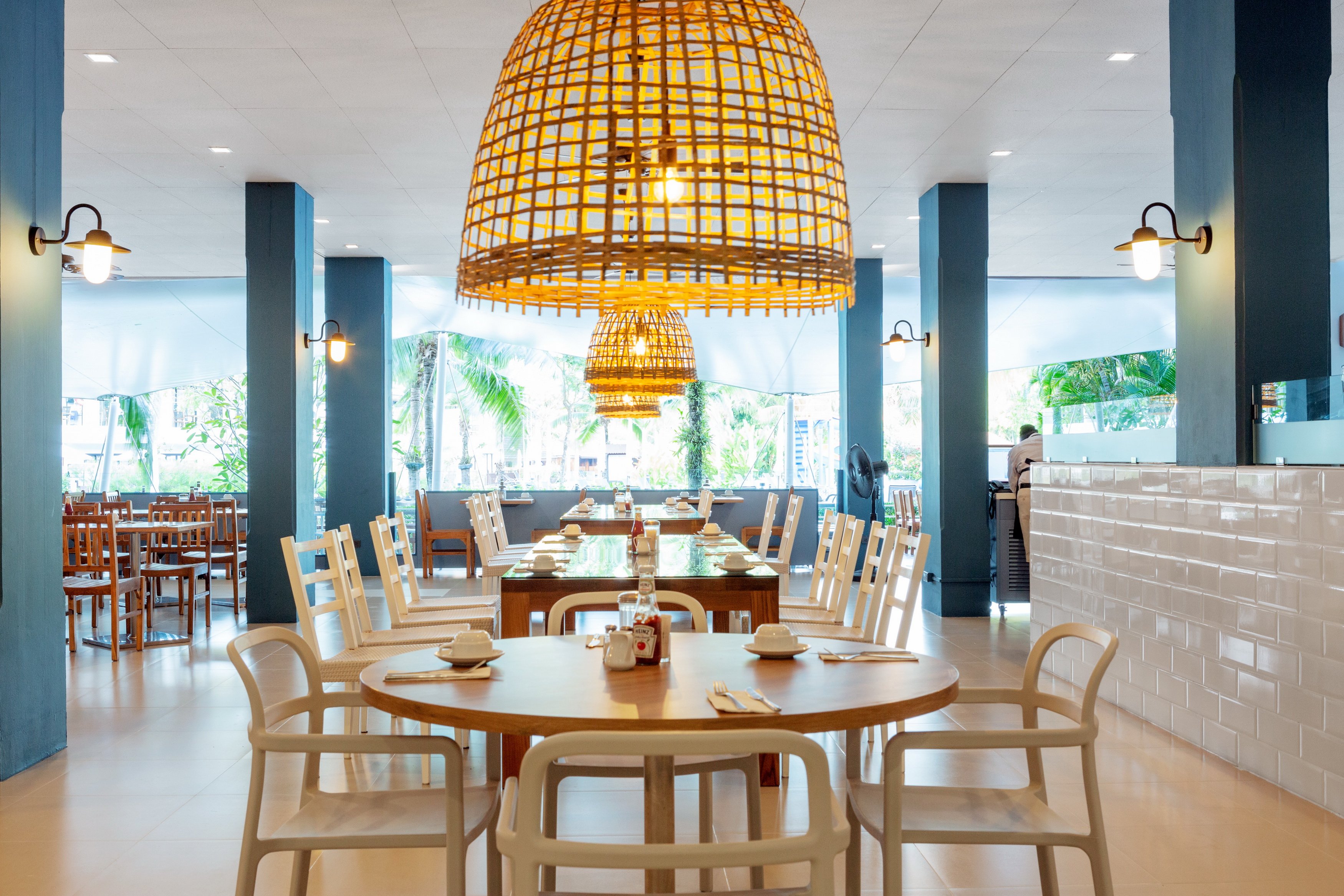 Sunwing Resort Kamala Beach restorāna iekštelpas ar modernām mēbelēm, lieliem pītiem gaismekļiem un skatu uz tropu dārzu.