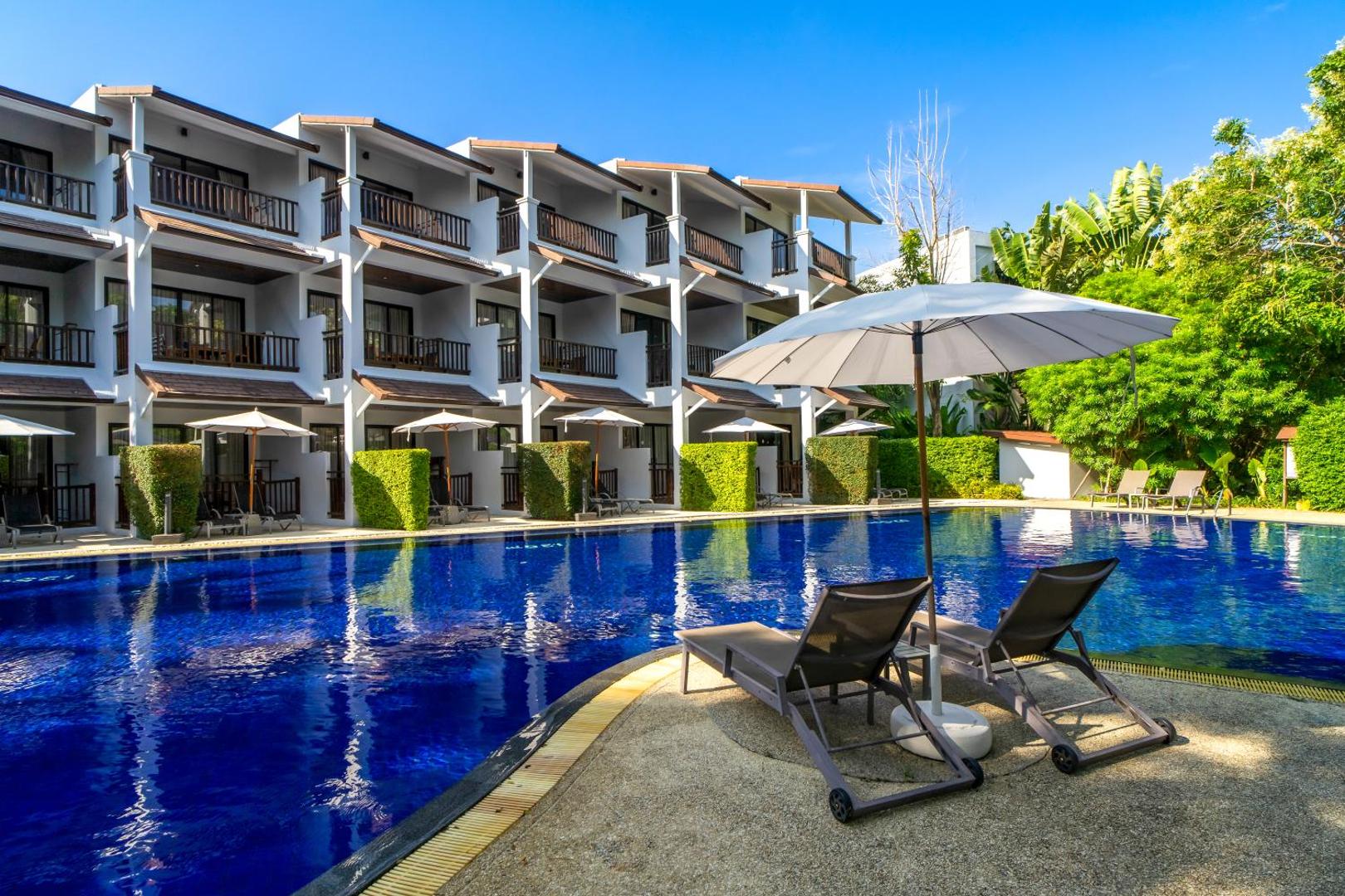 Atpūtas krēsli un saulessargs pie Sunwing Resort Kamala Beach lielā āra baseina — fonā redzami viesnīcas numuri ar balkoniem un tropu zaļumi