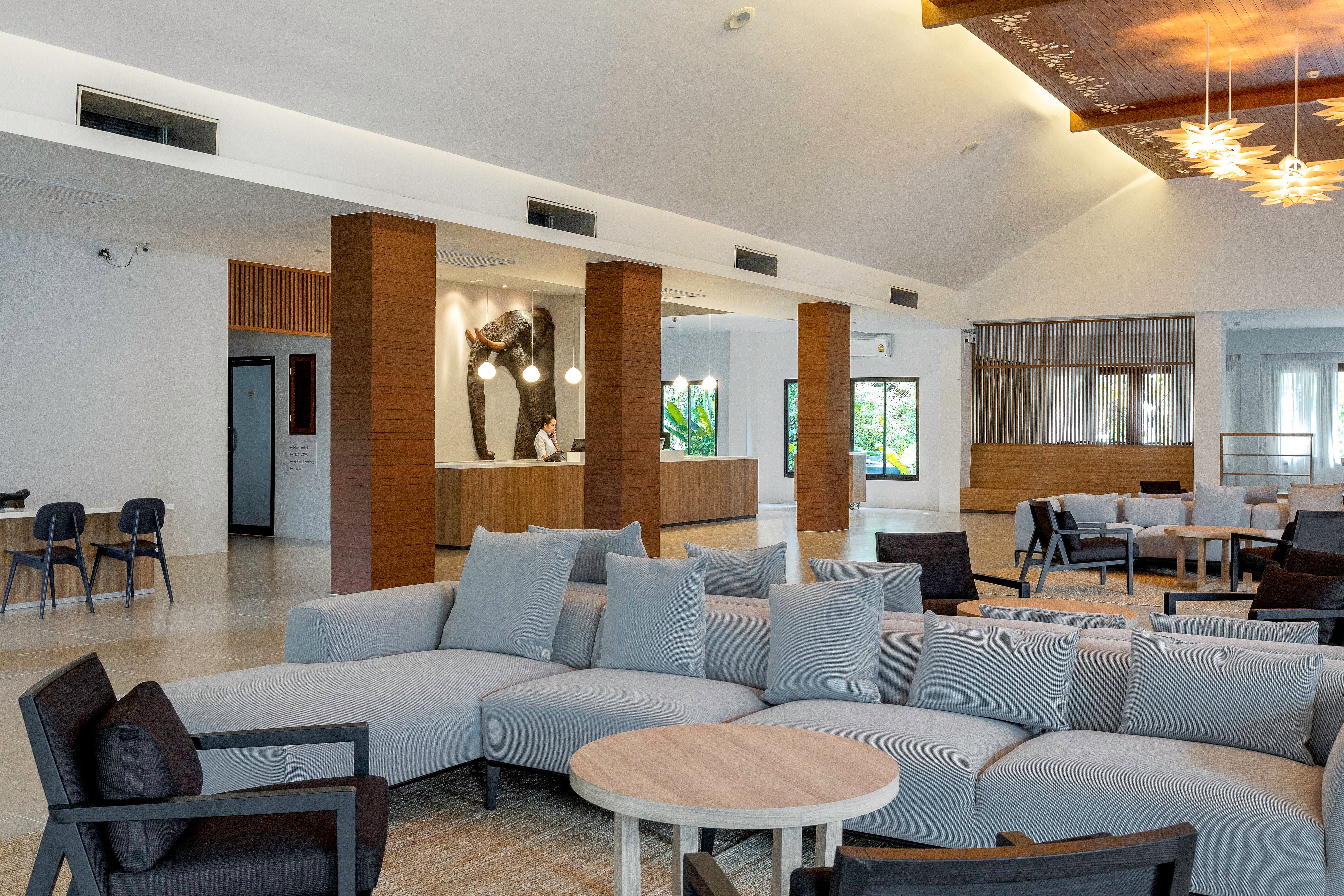 Sunwing Resort Kamala Beach plašā lobby zona ar moderniem pelēkiem dīvāniem, koka kolonnām un stilīgu interjeru Puketas kūrortā.