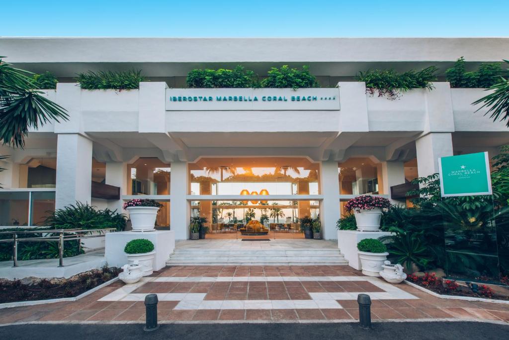 Iberostar Marbella Coral Beach viesnīcas ieeja ar modernu arhitektūru, zaļiem augiem un ziediem, kas rada relaksējošu atmosfēru. Elegantā fasāde un plašais ieejas laukums aicina viesus izbaudīt brīvdienas pie jūras.