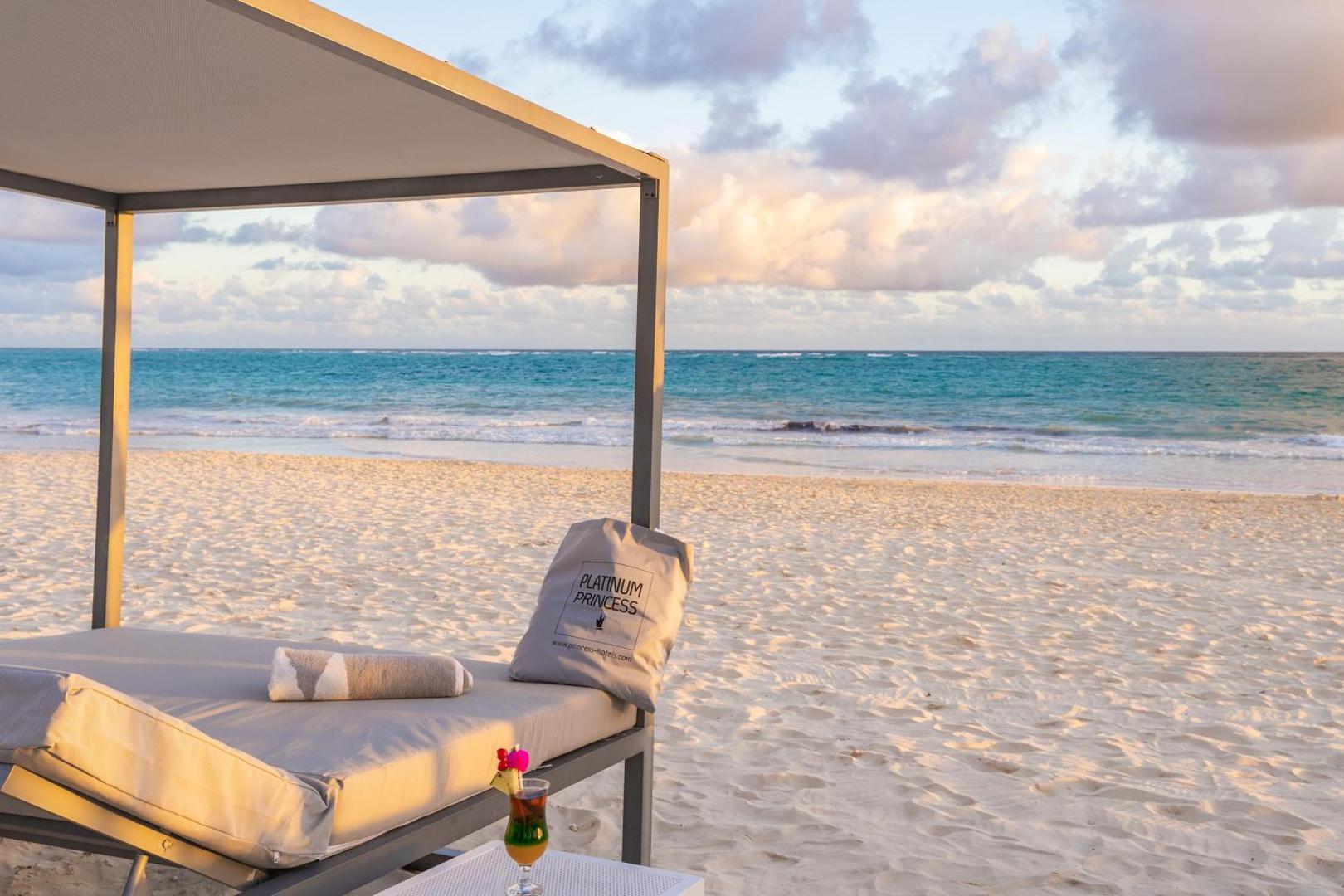 Grand Bavaro Princess privātā pludmales zona ar ērtu sauļošanās gultu, kokteili un skatu uz tirkīzzilo okeānu saulrietā