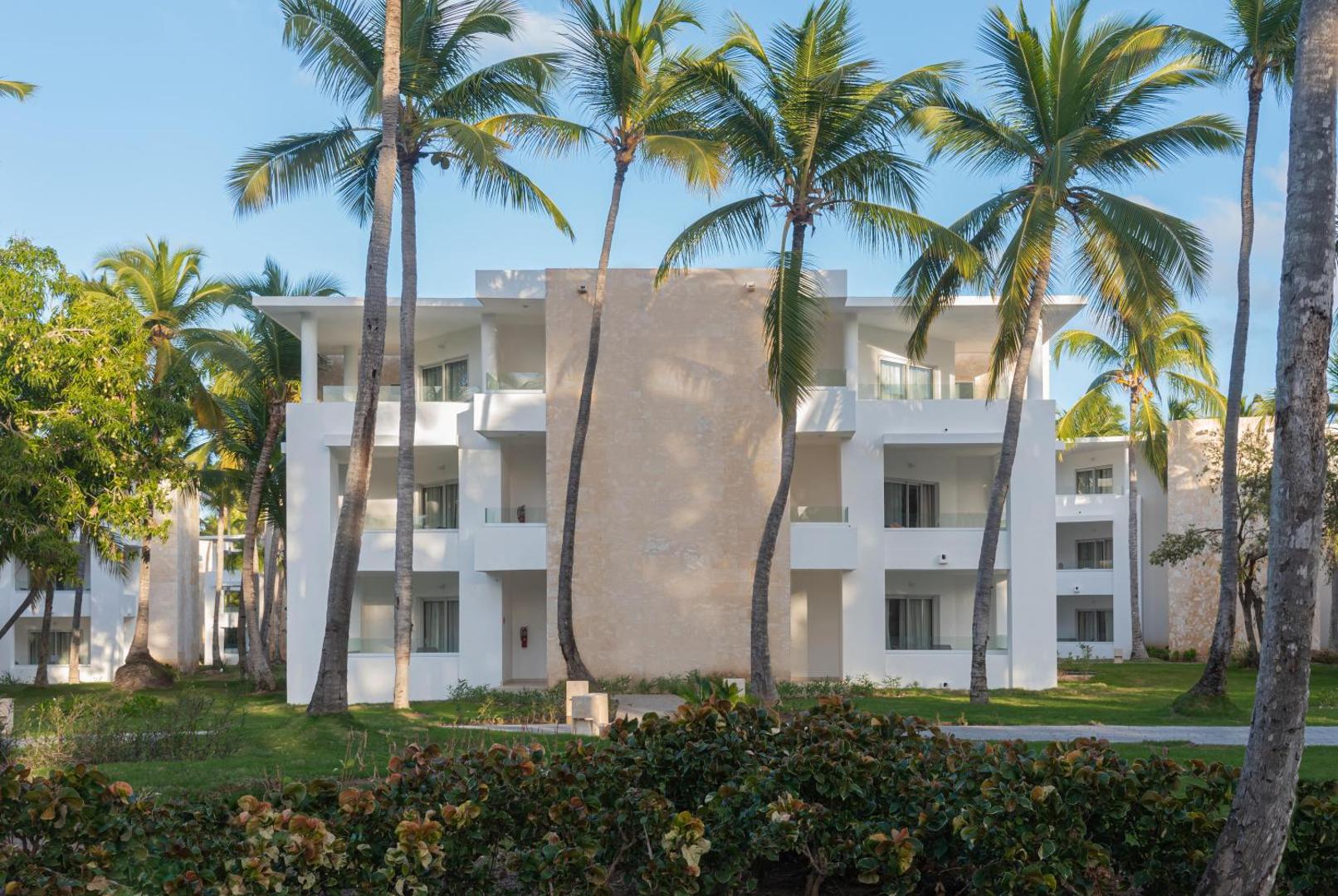 Grand Bavaro Princess trīsstāvu numuru korpuss ar balkoniem, ko ieskauj palmas un tropu apstādījumi saulainā dienā