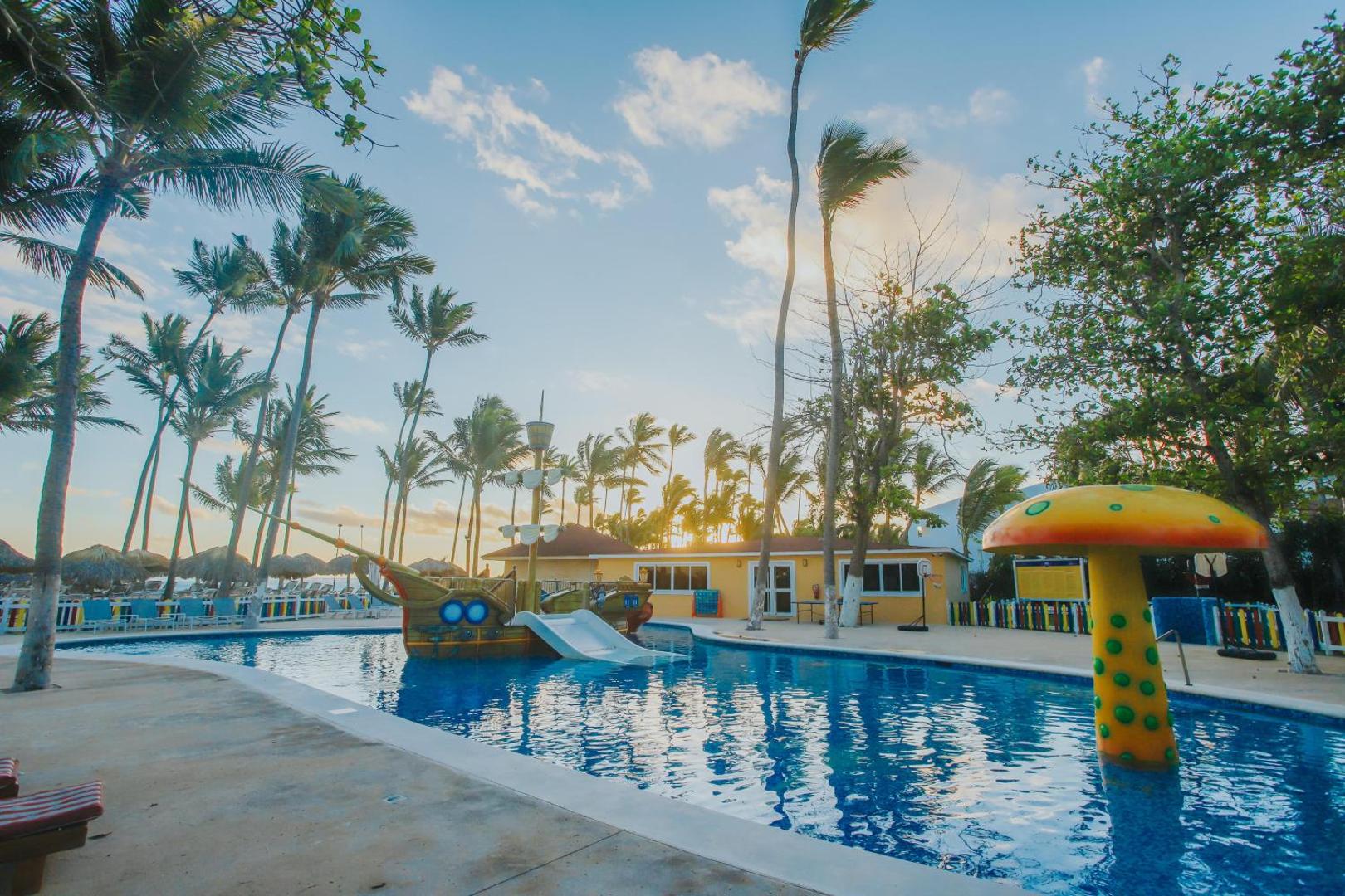 Grand Bavaro Princess bērnu baseins ar pirātu kuģi un slidkalniņu palmu ieskautā teritorijā Punta Kanā saulrieta laikā