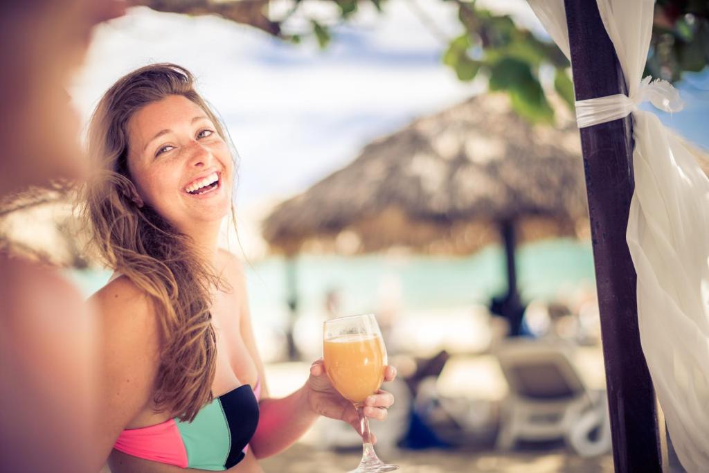 Smaidoša sieviete pludmalē, turot rokā dzērienu, ar fona palmām un pludmales gultām.