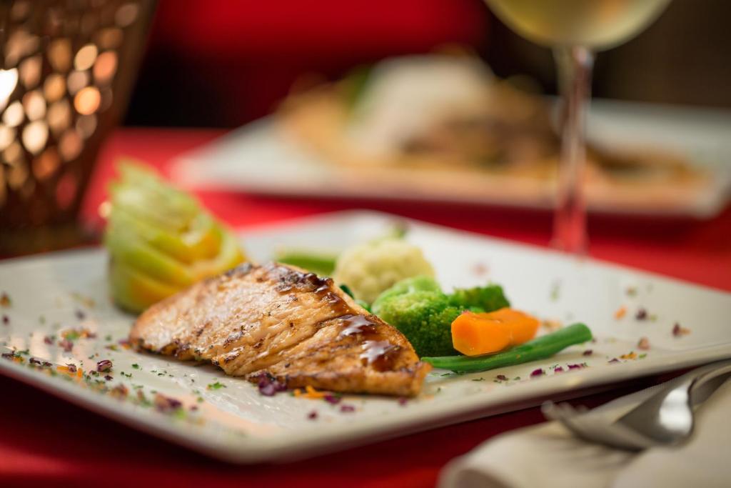 Garšīgi pasniegta cepta laša fileja ar dārzeņu garnējumu, ieskaitot brokoļus, burkānus un kartupeļu biezeni, uz eleganta galda ar sarkanā galdauta fonu.