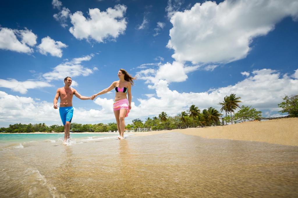 Pāris, turot rokas, staigā pa pludmali zem saulainām debesīm ar mākonīšiem. Apkārt ir palmas un mierīga jūra.