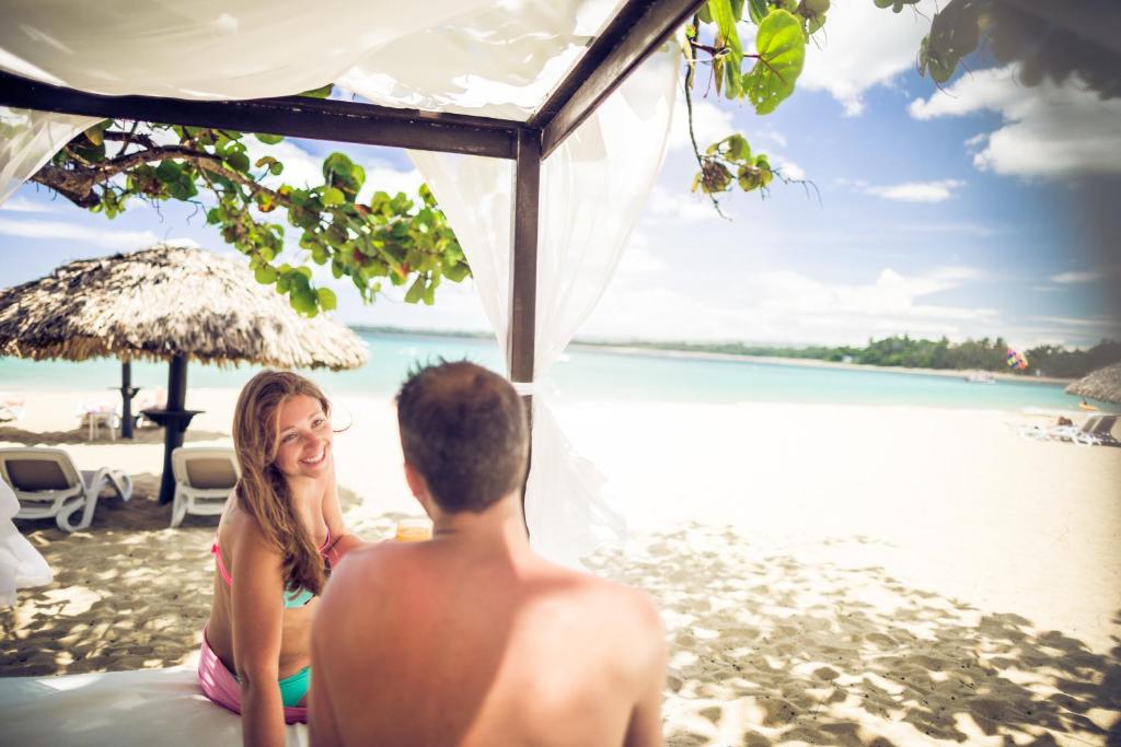 Pludmales ainava ar diviem cilvēkiem, kas sarunājas zem nojumes, fonā redzama zila jūra un saulaina debesis.