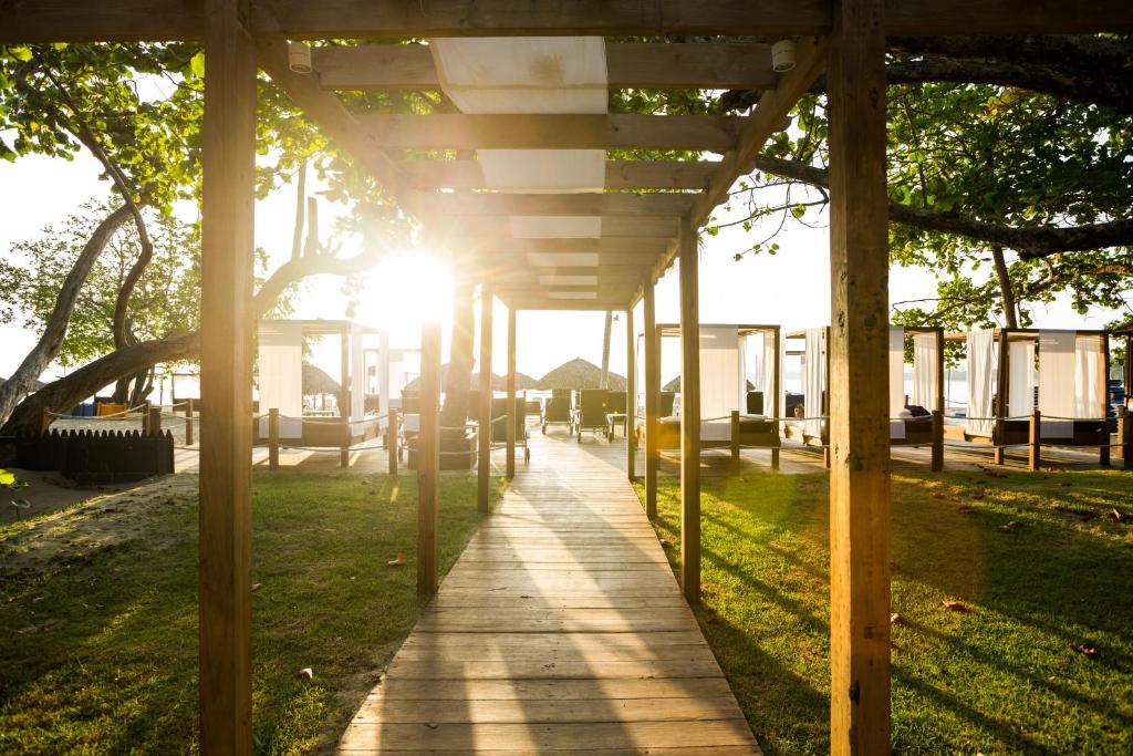 Saulaina promenāde, kas vijas cauri viesnīcas teritorijai, ar zāles un koka struktūrām, kas nodrošina relaksējošu atmosfēru. Gaisma caur koku lapām rada siltu un viesmīlīgu noskaņu.