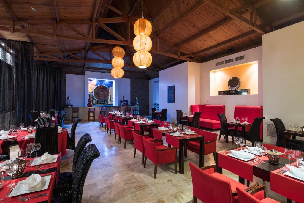 Mūsdienīgs restorāns ar sarkaniem krēsliem un elegantu galdu uzstādījumu, ko apgaismo liela, stilīga apgaismojuma ķermeņa. Interjers ar koka griestiem un melnām aizkariem rada siltu un viesmīlīgu atmosfēru.