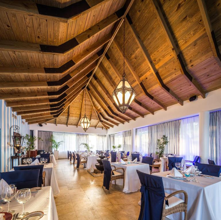 Elegants restorāna interjers ar koka griestiem un modernām lampām, kur galdi ir klāti ar baltām un tumši zilu galdautiem. Gaismas un augi piešķir telpai patīkamu atmosfēru.