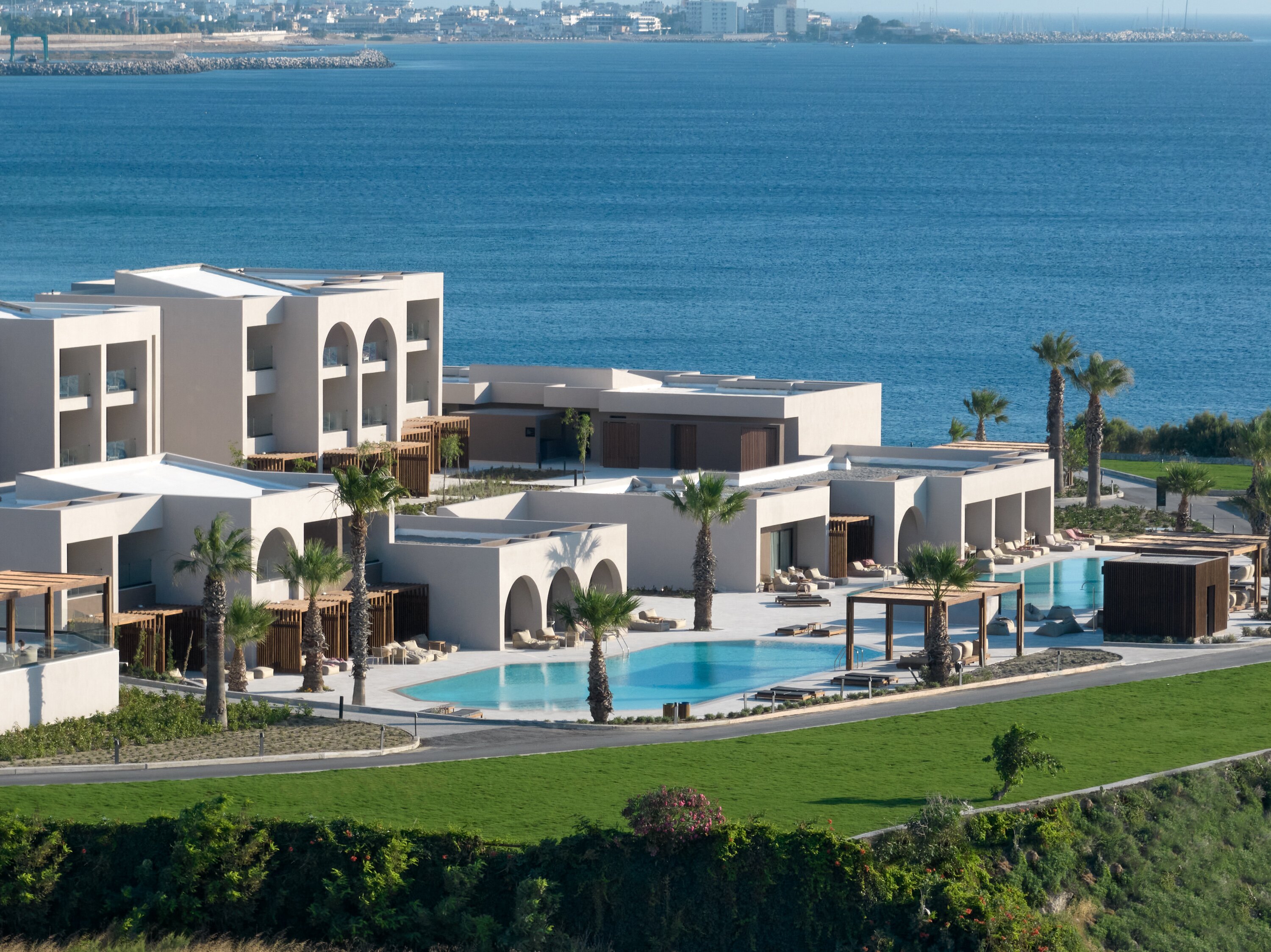 Moderns kūrortviesnīcas komplekss pie jūras, ar baseinu un palmām, kas rada relaksējošu atmosfēru. Apkārt ir zaļš zāliens un skats uz ūdeni. panoramic view with beach and pools
