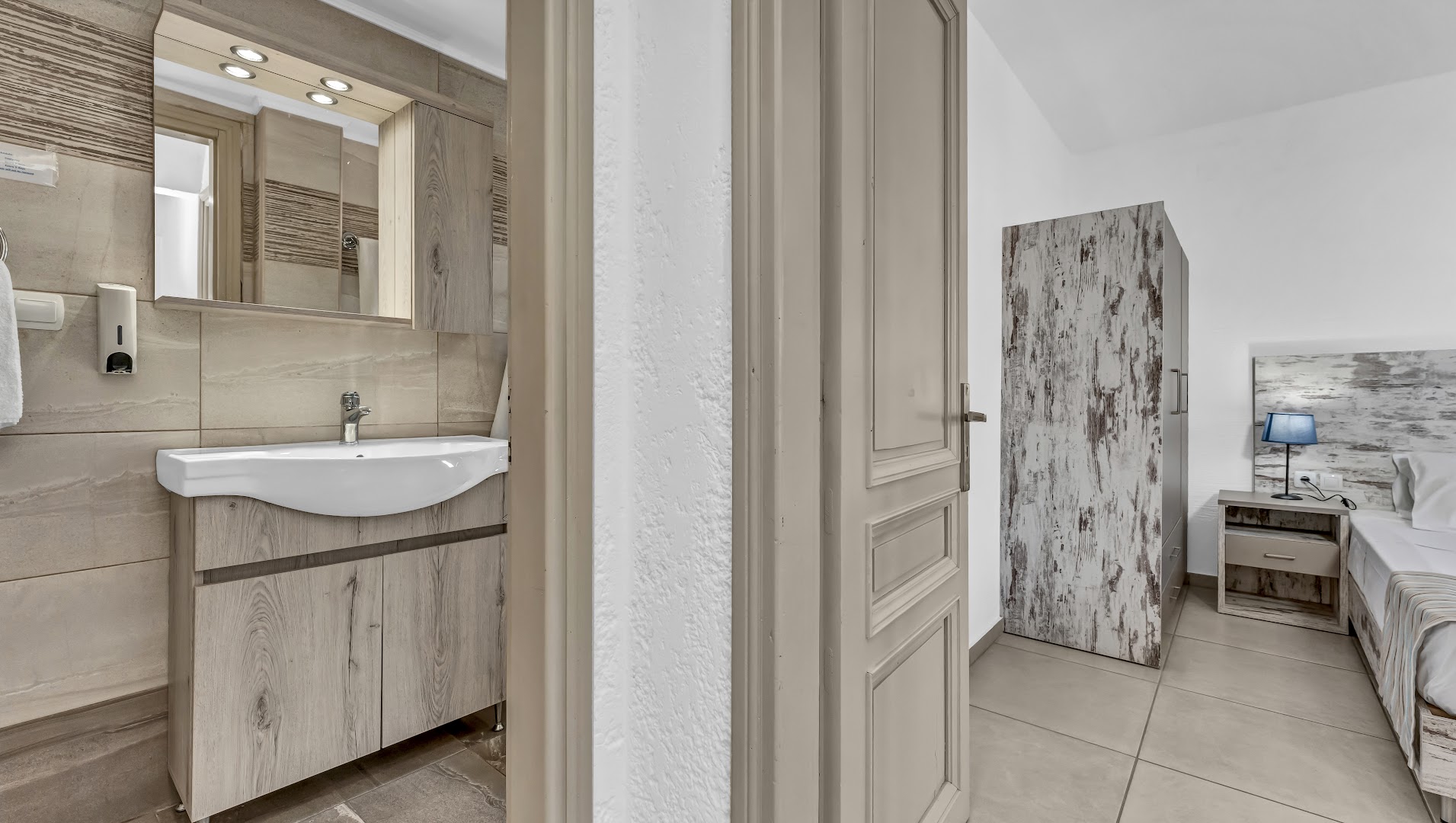 Evina Rooms & Suites gaišs numurs ar skatu uz vannas istabu un guļamzonu, kurā ir koka mēbeles un zila naktslampa