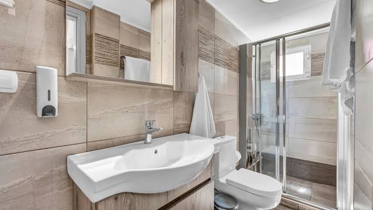 Evina Rooms & Suites modernā vannas istaba ar dušas kabīni, gaišām flīzēm un koka apdares elementiem viesnīcas numurā