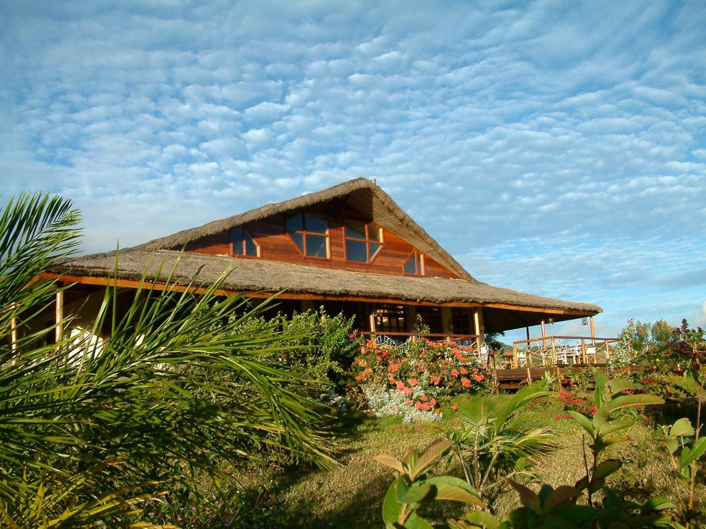 Nature Lodge galvenā ēka ar salmu jumtu un ziedošu dārzu saulainā dienā.
