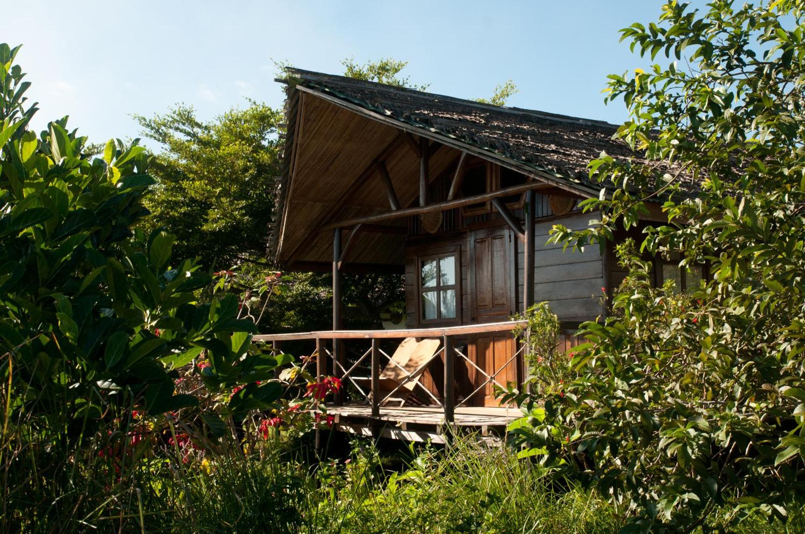 Mājīgs koka bungalo ar atpūtas krēsliem uz terases starp zaļiem kokiem.