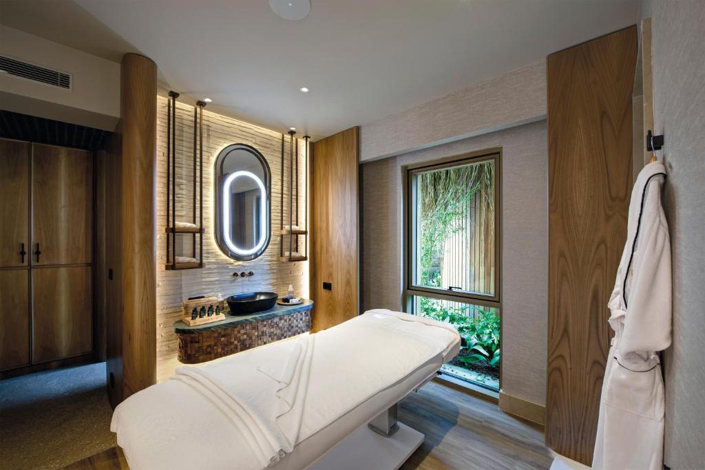 Mūsdienīgs spa telpas interjers ar masāžas galdu, elegantiem koka akcentiem un dabu caur logu. Telpa nodrošina relaksējošu atmosfēru ar mūsdienīgu dizainu un ērtībām.