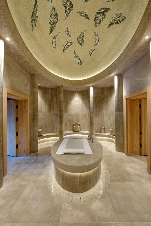 Mūsdienīgs spa interjers ar elegantu apgaismojumu un dabīgu akmeņu elementiem, kas rada relaksējošu atmosfēru. Uz griestiem redzami zaļi lapu motīvi, piešķirot telpai dzīvīgumu.