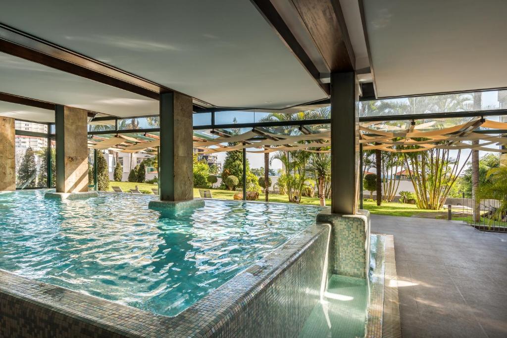 Mūsdienīgs iekštelpu baseins ar stikla sienām un dabīgu apgaismojumu, kas sniedzas uz zāli, kur redzami zaļie augi un sauļošanās krēsli. Ideāla vieta relaksācijai un atpūtai.