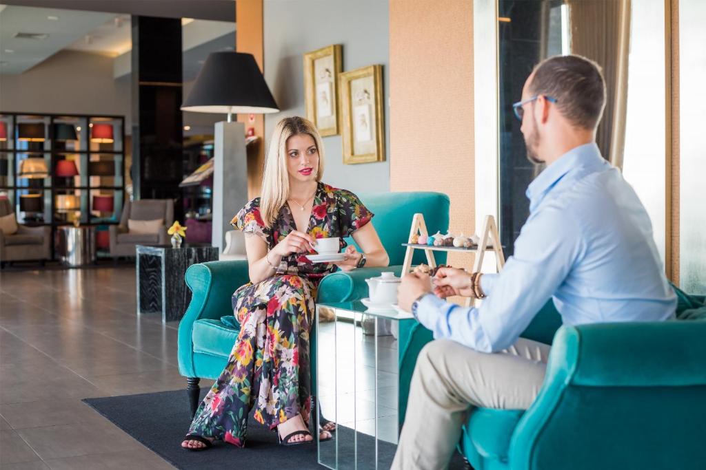 Sieviete ar ziedu apdruku tērpā sēž pie galda, turot krūzi, kamēr sarunājas ar vīrieti. Abiem ir moderni apģērbi, un fonā ir luksusa viesnīcas interjers ar stilīgām mēbelēm.