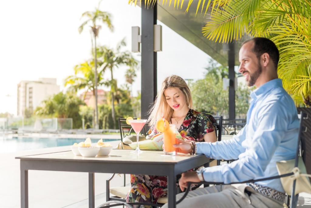 Pāris relaksējas, baudot dzērienus un ēdienus terasē pie baseina, ar palmām un saulainu atmosfēru apkārt.