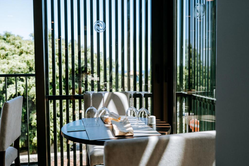 Mūsdienīga restorāna interjers ar apaļu galdu, uz kura ir glāzes un galda piederumi, caur stikla durvīm ieplūst dabiska gaisma. Fonā redzama zaļā ainava.